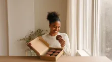 A smiling woman sits by a sunlit window, holding a chocolate cookie from an open box filled with cookies, creating a warm and inviting scene that highlights mindful enjoyment of food.